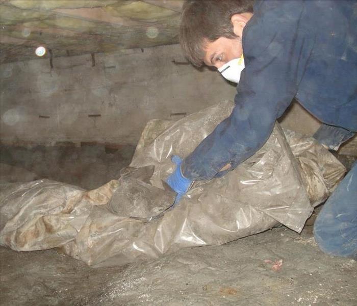 Man with blue overalls and safety mask on in crawl space rolling up plastic drop cloth 